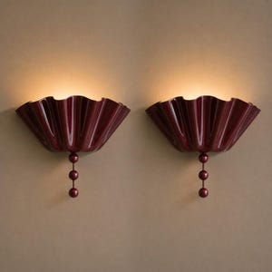 Peut inclure: Une paire d'appliques murales rouge foncé dotées d'un large abat-jour plissé en forme d'éventail avec une finition brillante. Chaque applique est éclairée de l'intérieur et comporte un élément décoratif vertical composé de trois petites sphères.