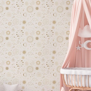 Op de afbeelding: Een kinderkamerscène met een lichtroze luifel over een wieg van riet. Het behang heeft een hemels patroon van gouden zonnen, manen en sterren. Een geweven vloerkleed en decoratieve items maken de inrichting van de kamer compleet.