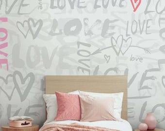 May include: A bedroom with a white wall covered in gray and pink graffiti that says "love". The bed has a pink blanket and two pink pillows. There is a small wooden table with a pink box on it and a vase with a pink flower on a wooden table next to the bed.