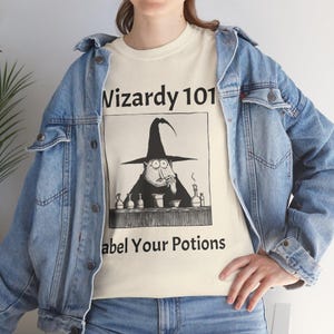 May include: Cream-colored t-shirt with a black and white cartoon of a wizard with the text "Wizardy 101" and "Label Your Potions". The wizard is wearing a tall hat and is surrounded by potion bottles.