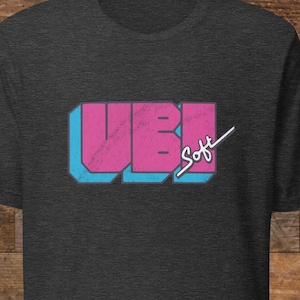 Op de afbeelding: Donkergrijs t-shirt met een retro design. Het ontwerp toont de letters "UBI" in roze met een blauwe omlijning en het woord "Soft" in witte letters. Het shirt is gemaakt van een zacht materiaal.