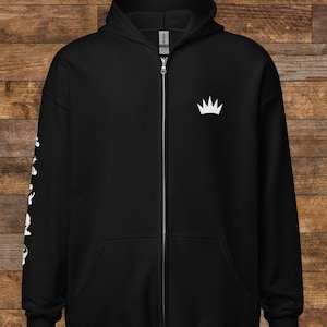 May include: Black zip-up hoodie featuring a white crown logo on the chest and white graphics on the left sleeve. The hoodie has a drawstring hood and a front pocket. The background is a wooden surface.