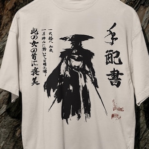 Puede incluir: Camiseta de color crema con una ilustración en tinta negra de un guerrero samurái y caligrafía japonesa. El texto incluye la palabra "Se busca" y otras frases. La camiseta está colgada en una percha de madera sobre un fondo texturizado.