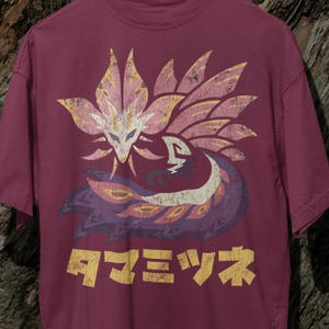 May include: Burgundy t-shirt with a graphic of a mythical creature with floral elements in pink, yellow, and purple. Japanese text in yellow is visible. The shirt is displayed on a wooden hanger.