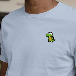 Könnte beinhalten: Hellblaues T-Shirt mit einem pixeligen grünen Alligator-Design auf der Brust. Das kurzärmelige T-Shirt ist aus einem weichen Material gefertigt und eignet sich für Freizeitkleidung. Das Alligator-Design ist in Grün-, Gelb- und Brauntönen gehalten.