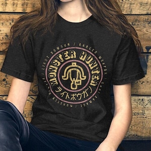 May include: A black t-shirt with a gold and pink circle graphic featuring the text "Monster Hunter" and Japanese characters. The graphic also includes a stylized image of a weapon.