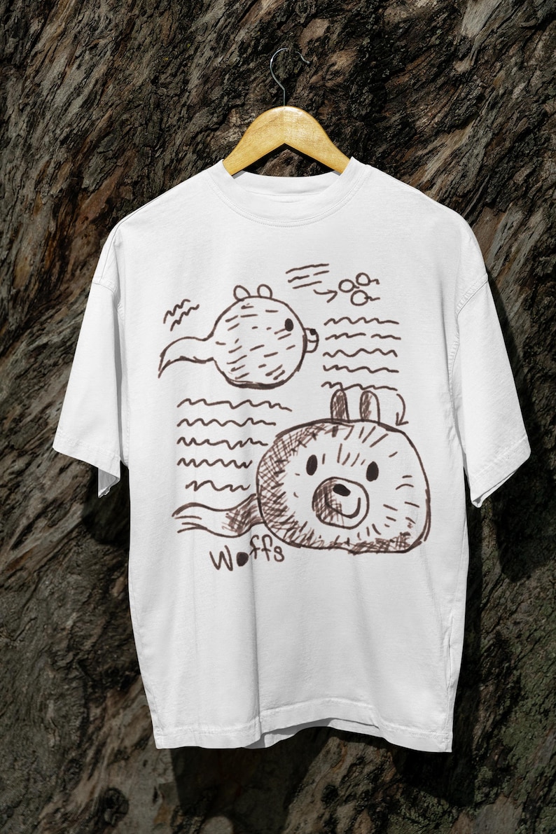 K&ouml;nnte beinhalten: Wei&szlig;es T-Shirt mit einer braunen Cartoon-Illustration von zwei runden Kreaturen und Wellenlinien. Die untere Kreatur tr&auml;gt das Wort "Woffs" darunter. Das T-Shirt h&auml;ngt an einem Holzb&uuml;gel.