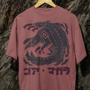 Puede incluir: Una camiseta de color marrón rojizo con un gráfico negro y morado de un monstruo con el texto japonés "コフ・キザク" impreso en la parte delantera.