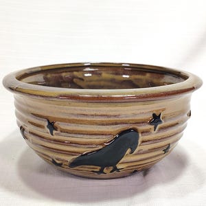 May include: A ceramic bowl with a brown and tan glaze. The bowl has horizontal ridges and is decorated with a black raven and star cutouts. The rim is a darker brown. The bowl is likely handmade and suitable for decorative or functional use.
