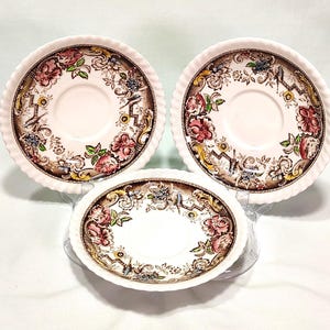 May include: Three pieces of vintage china with a floral design. The set includes two round saucers and one oval dish. The rims have a white, ridged texture. The floral pattern features red, pink, and yellow flowers with green leaves on a brown background.