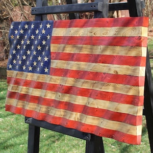 May include: A wooden American flag with a distressed finish, displayed on a black easel. The flag features red and natural wood stripes, a blue field with gold stars, and a wavy design. The artwork is displayed outdoors.