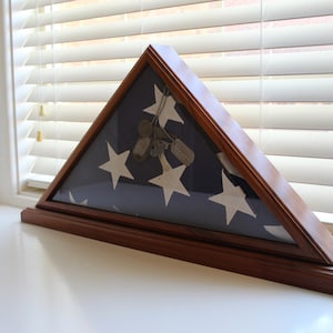 May include: Triangular wooden flag display case with a glass front. The case holds a folded American flag with white stars on a blue field. Inside the case are several dog tags. The case is sitting on a white surface.