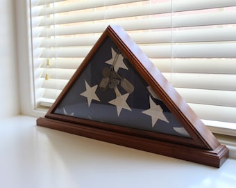 May include: Triangular wooden flag display case with a glass front. The case holds a folded American flag with white stars on a blue field. Inside the case are several dog tags. The case is sitting on a white surface.