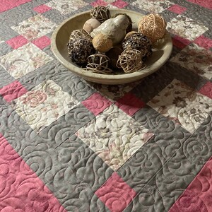 Könnte beinhalten: Ein gesteppter Tischläufer mit einem Blumenmuster in cremefarbenen und rosa Quadraten, umrandet von grauem und rosa Stoff. Eine Holzschale mit dekorativen Kugeln und einem Keramikvogel liegt auf dem Läufer.