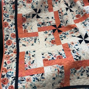 Könnte beinhalten: Ein floraler Quilt mit einem Patchwork-Design. Der Quilt hat einen weißen Hintergrund mit einem Muster aus Aquarellrosen in Pfirsich-, Blau- und Weißtönen. Der Quilt hat einen marineblauen Rand und pfirsichfarbene Akzente. Der Quilt ist über einer dunklen Oberfläche gefaltet.