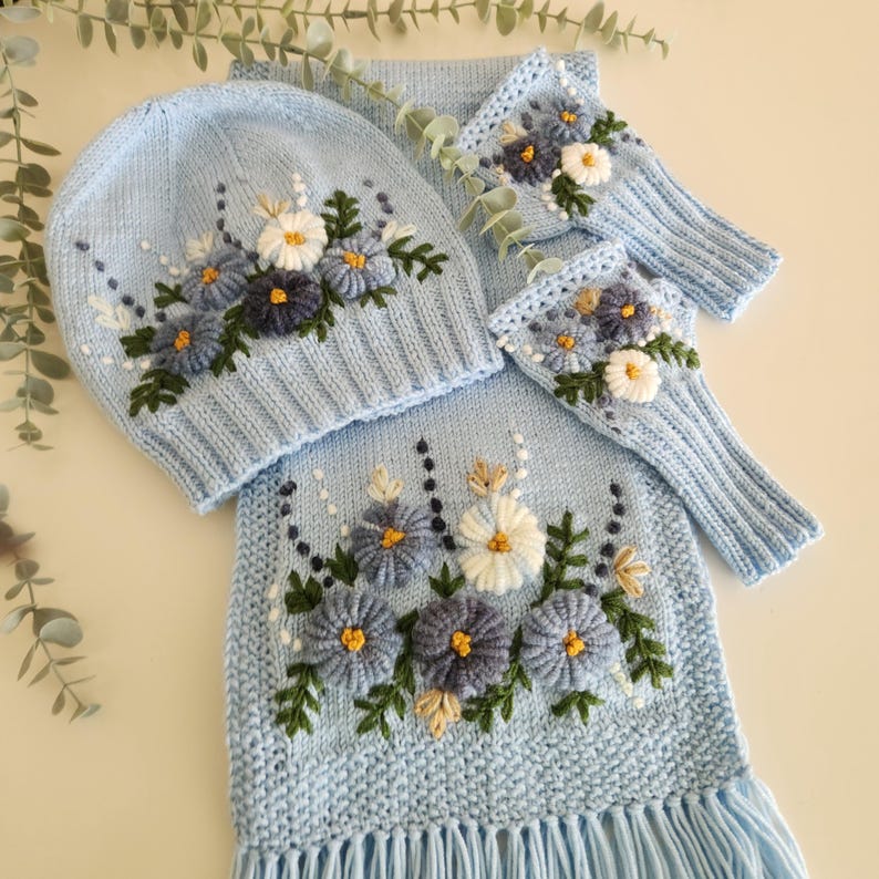 May include: A light blue knit hat, scarf, and fingerless gloves set. The set features embroidered flowers in shades of blue, white, and yellow, with green leaves. The scarf has a fringe at the bottom. The set is designed for cold weather.