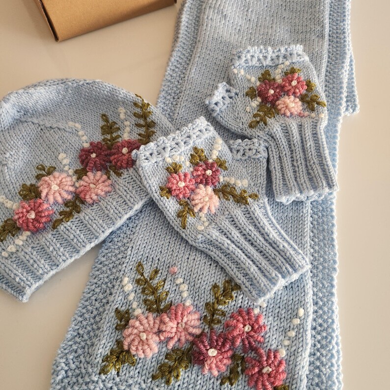 May include: A light blue knitted set featuring a hat, scarf, and fingerless gloves, all adorned with embroidered pink and burgundy flowers, green leaves, and white accents. The set is made of soft, textured yarn.