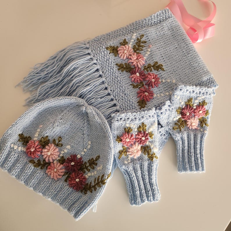 May include: A light blue knitted hat, scarf, and fingerless gloves set. The set features embroidered pink and burgundy flowers with green leaves. The scarf has a fringe, and the set is displayed on a white surface.