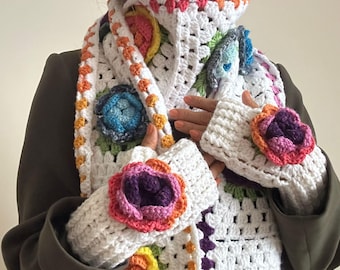 May include: A white crochet scarf and matching wrist warmers with colorful floral accents. The scarf and wrist warmers feature a granny square design with vibrant, multi-colored flowers in shades of pink, purple, blue, and yellow. The edges are trimmed with orange and pink.