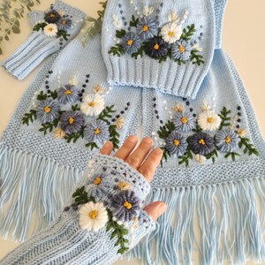 May include: A light blue knitted set featuring a scarf with fringe, a hat, and fingerless gloves, all adorned with embroidered blue, white, and yellow flowers and green leaves. The set is made of soft, textured yarn.