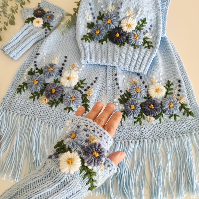 May include: A light blue knitted set featuring a scarf with fringe, a hat, and fingerless gloves, all adorned with embroidered blue, white, and yellow flowers and green leaves. The set is made of soft, textured yarn.