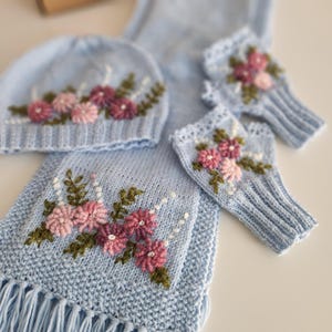 May include: A light blue knitted set featuring a hat, scarf, and fingerless gloves, all adorned with embroidered pink and purple flowers and green leaves. The scarf has a fringe. A cozy, handmade winter accessory.