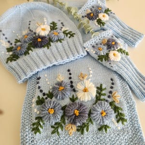 May include: A light blue knit hat, scarf, and fingerless gloves set. The set features embroidered flowers in white, blue, and yellow with green leaves. The items are arranged on a white surface.