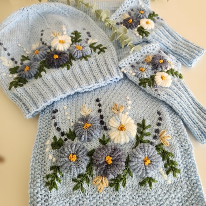 May include: A light blue knit hat, scarf, and fingerless gloves set. The set features embroidered flowers in white, blue, and yellow with green leaves. The items are arranged on a white surface.