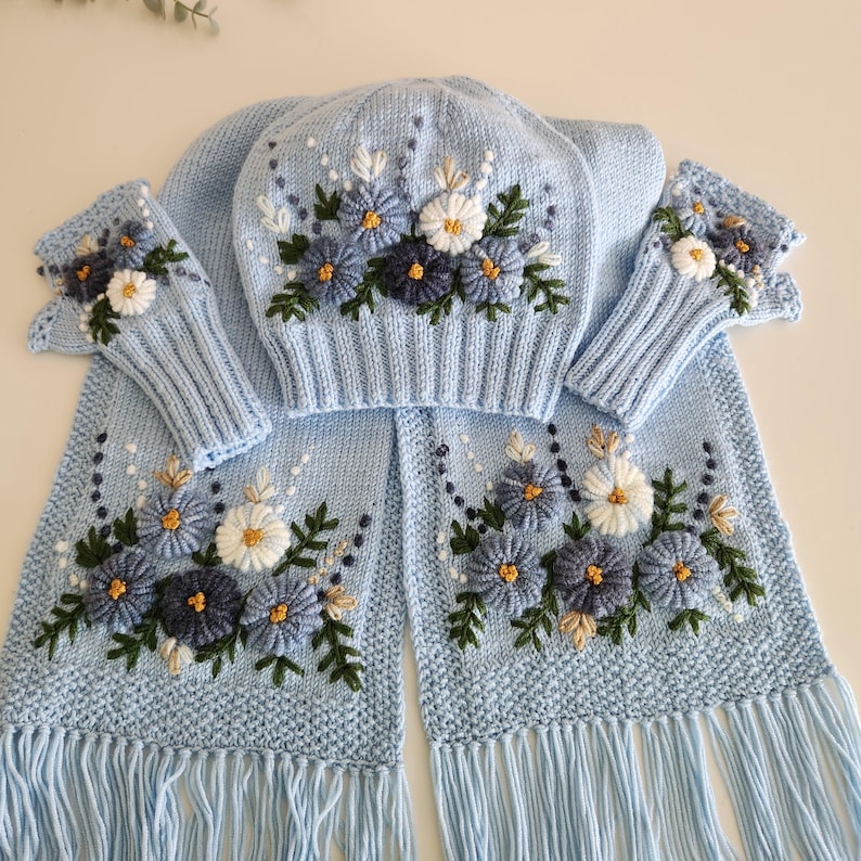 May include: A light blue knitted winter set featuring a hat, scarf, and fingerless gloves. The set is adorned with embroidered flowers in shades of blue, white, and yellow. The scarf has a fringed edge, and the gloves are open at the fingers.