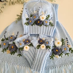 May include: A light blue knitted winter set featuring a hat, scarf, and fingerless gloves, all adorned with embroidered floral designs in shades of blue, white, and yellow. The scarf has a fringed edge.