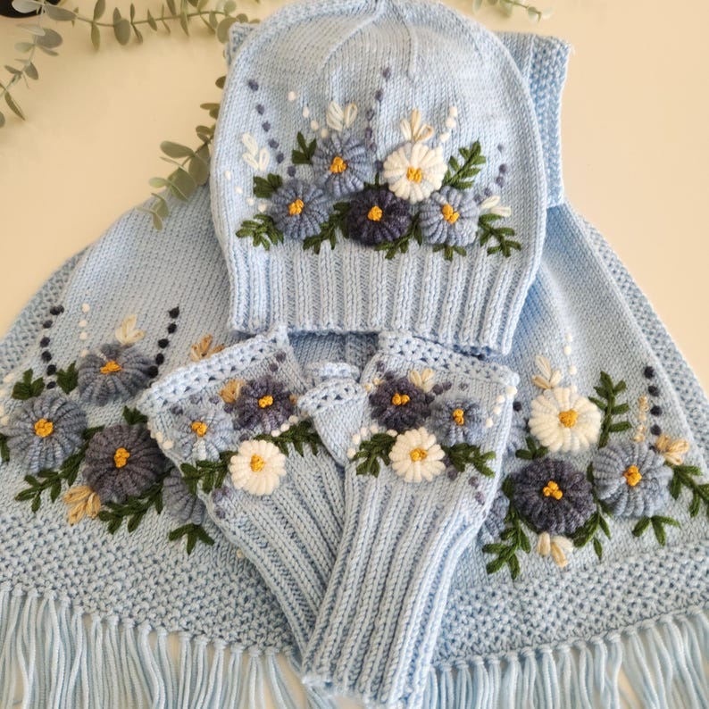 May include: A light blue knitted winter set featuring a hat, scarf, and fingerless gloves, all adorned with embroidered floral designs in shades of blue, white, and yellow. The scarf has a fringed edge.