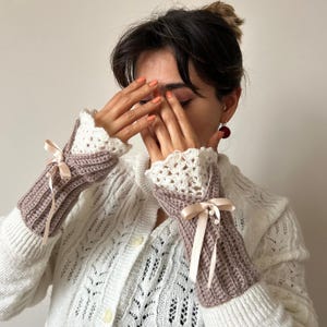 May include: Crocheted fingerless gloves in mauve and white, featuring a ribbed design and delicate lace trim. The gloves are adorned with satin ribbons and small pearl accents, adding a touch of elegance. The overall style is vintage-inspired and handmade.