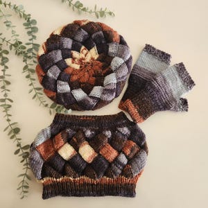 May include: A knitted set in brown, grey, and cream, comprising a beret, fingerless gloves, and a cowl. The beret has a woven design with a central flower-like pattern. The fingerless gloves and cowl have a similar woven texture.