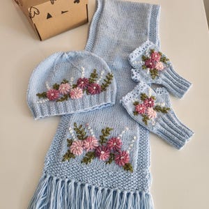 May include: A light blue knitted hat, scarf, and fingerless gloves set. Each piece is adorned with embroidered pink and purple flowers with green leaves. The scarf has a fringed edge. The set is displayed on a white surface.