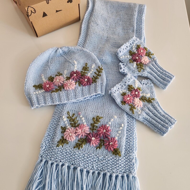 May include: A light blue knitted hat, scarf, and fingerless gloves set. Each piece is adorned with embroidered pink and purple flowers with green leaves. The scarf has a fringed edge. The set is displayed on a white surface.