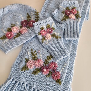 May include: A light blue knitted winter set featuring a hat, scarf, and fingerless gloves. The set is adorned with embroidered pink and burgundy flowers, green leaves, and white beaded accents. The scarf has a fringed edge.