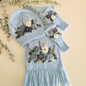 May include: A light blue knit hat, scarf, and fingerless gloves set. The set is adorned with embroidered flowers in white, blue, and yellow. The scarf has a fringed edge, and the set is displayed on a light background.