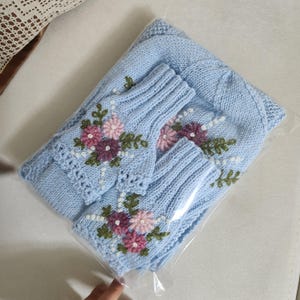 May include: A light blue knitted set, including a hat and fingerless gloves, is displayed in a clear plastic bag. The set is adorned with embroidered flowers in shades of pink and purple, with green leaves and white accents. The set is made of a soft, knitted material.