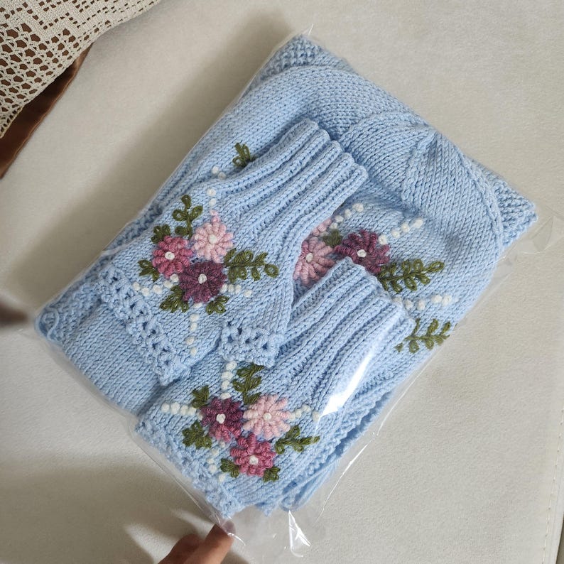 May include: A light blue knitted set, including a hat and fingerless gloves, is displayed in a clear plastic bag. The set is adorned with embroidered flowers in shades of pink and purple, with green leaves and white accents. The set is made of a soft, knitted material.