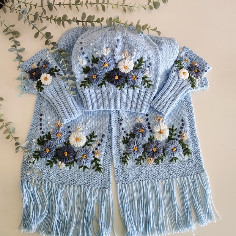 May include: A light blue knitted winter set featuring a hat, scarf, and fingerless gloves, all adorned with embroidered floral designs in shades of blue, white, and green. The scarf has a fringed edge.