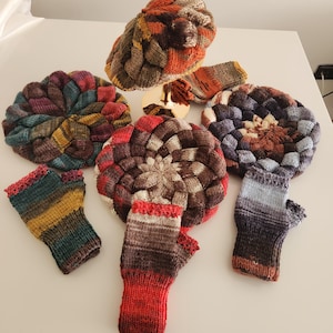 May include: A selection of hand-knitted hats and fingerless gloves. The hats have a textured, woven design in shades of brown, orange, and blue. The fingerless gloves feature a striped pattern with red trim. The items are displayed on a white surface.