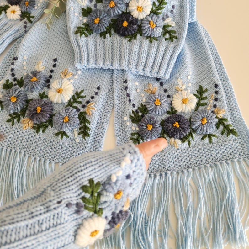 May include: A light blue knitted scarf, sweater, and fingerless gloves set. The set features embroidered flowers in shades of blue, white, and yellow. The scarf has a fringe along the bottom. The sweater is a crew neck. The gloves are fingerless.
