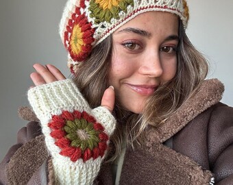 May include: A cream-colored crochet hat and fingerless gloves set with a floral design in red, yellow, and green. The hat is a beret style, and the gloves have a matching flower on the back of the hand. The set is worn with a brown jacket.