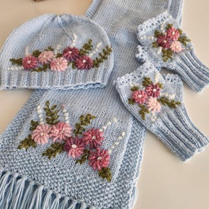 May include: A light blue knitted hat, scarf, and fingerless gloves set. The set is adorned with embroidered pink and purple flowers with green leaves. The scarf has a fringed edge. The set is displayed on a white surface.