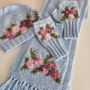 May include: A light blue knitted winter set featuring a hat, scarf, and fingerless gloves. The set is adorned with embroidered pink and burgundy flowers, green leaves, and white beaded accents. The scarf has a fringed edge.
