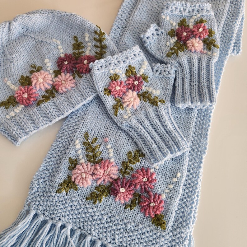 May include: A light blue knitted winter set featuring a hat, scarf, and fingerless gloves. The set is adorned with embroidered pink and burgundy flowers, green leaves, and white beaded accents. The scarf has a fringed edge.