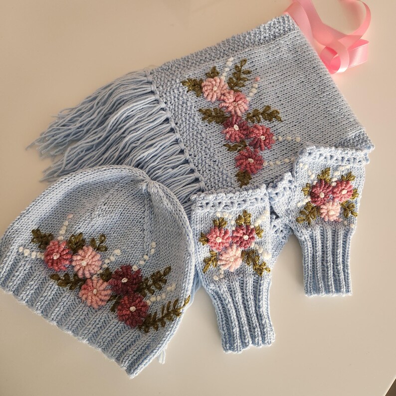 May include: A light blue knitted hat, scarf, and fingerless gloves set. The set features embroidered pink and burgundy flowers with green leaves. The scarf has fringe and a pink ribbon. The hat and gloves have a matching floral design.