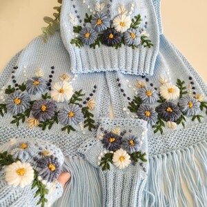 May include: A light blue knitted set featuring floral embroidery. The set includes a hat, scarf with fringe, and fingerless gloves. The flowers are white, blue, and dark blue with yellow centers and green leaves. The knitted items are made of soft yarn.