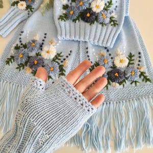 May include: Light blue knitted set featuring floral embroidery. The set includes a sweater, a scarf with fringe, and fingerless gloves. The flowers are white, blue, and navy with yellow centers and green leaves. The gloves are ribbed.