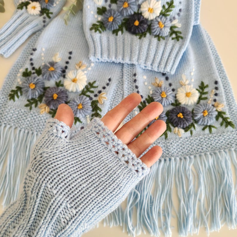 May include: Light blue knitted set featuring floral embroidery. The set includes a sweater, a scarf with fringe, and fingerless gloves. The flowers are white, blue, and navy with yellow centers and green leaves. The gloves are ribbed.